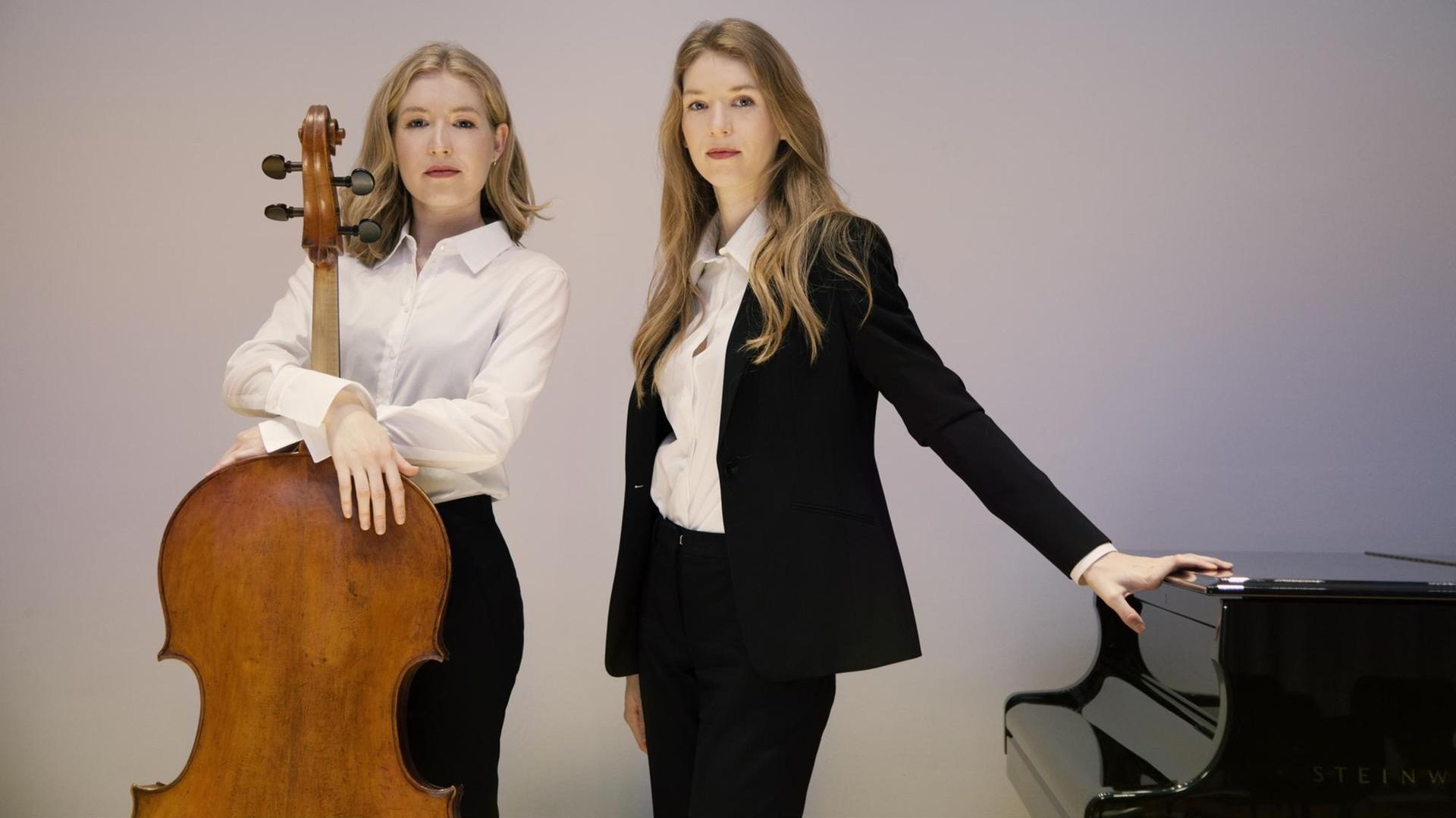Zwei junge Frauen stehen nebeneinander und schauen in die Kamera. Die linke hält ihr Cello vor sich, die rechte hat ihre Hand auf einem geschlossenen Flügel abgelegt. Zwei junge Frauen stehen nebeneinander und schauen in die Kamera. Die linke hält ihr Cello vor sich, die rechte hat ihre Hand auf einem geschlossenen Flügel abgelegt.