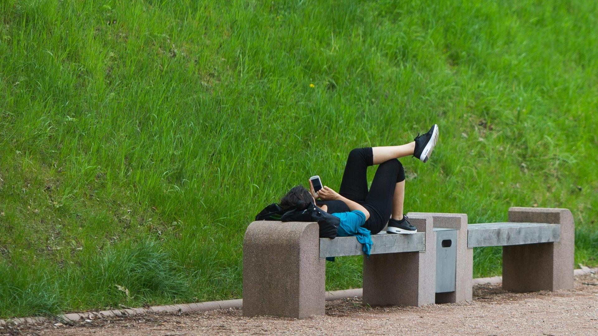 Eine Frau liegt auf einer Steinbank und schaut dabei auf ihr Telefon