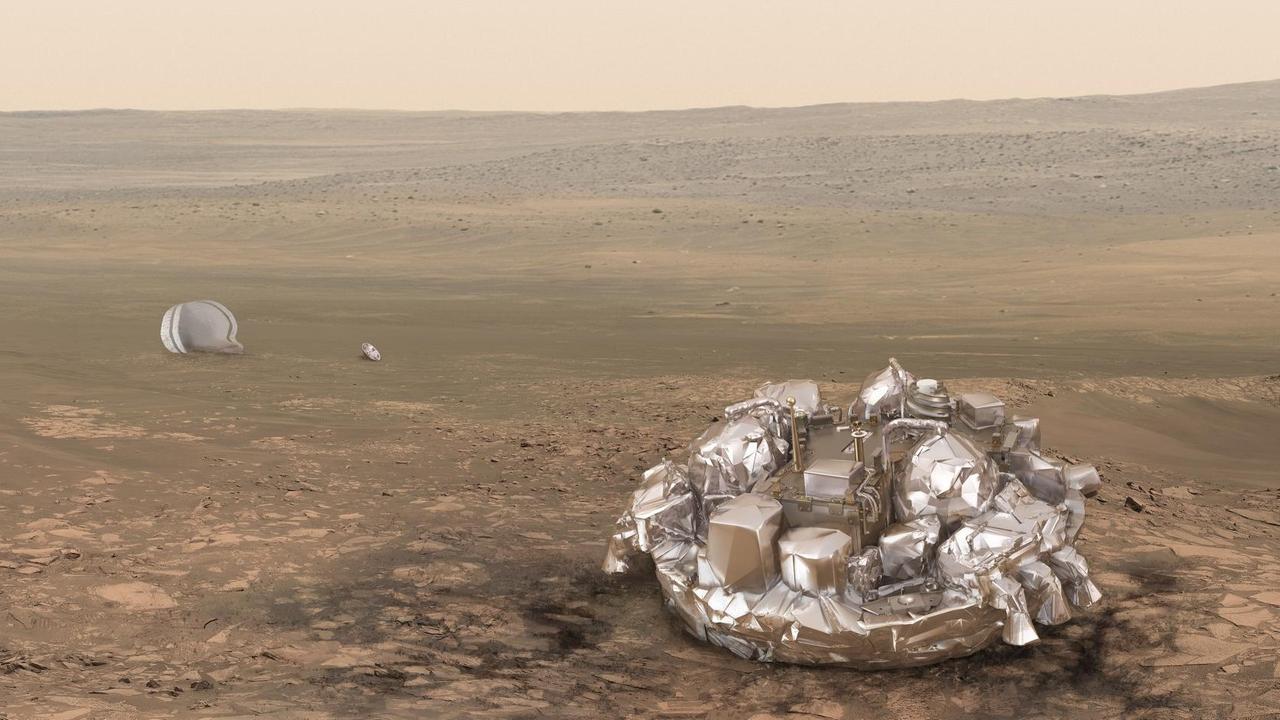 Die mit silberner Folie verkleidete Sonde liegt auf dem wüstenähnlichen Mars-Boden, im Hintergrund liegt der Fallschirm.