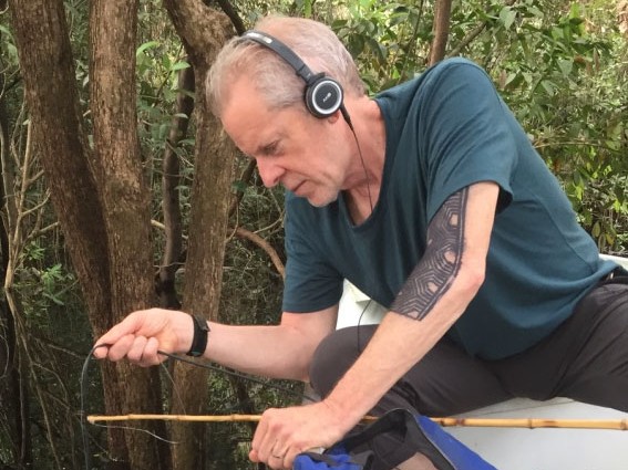 Robin Minard sitzt am Bootsrand, hält eine Schnur ins Wasser und hört über einem Kopfhörer Unterwasserklänge im Regenwald, Amazonas Robin Minard sitzt am Bootsrand, hält eine Schnur ins Wasser und hört über einem Kopfhörer Unterwasserklänge im Regenwald, Amazonas