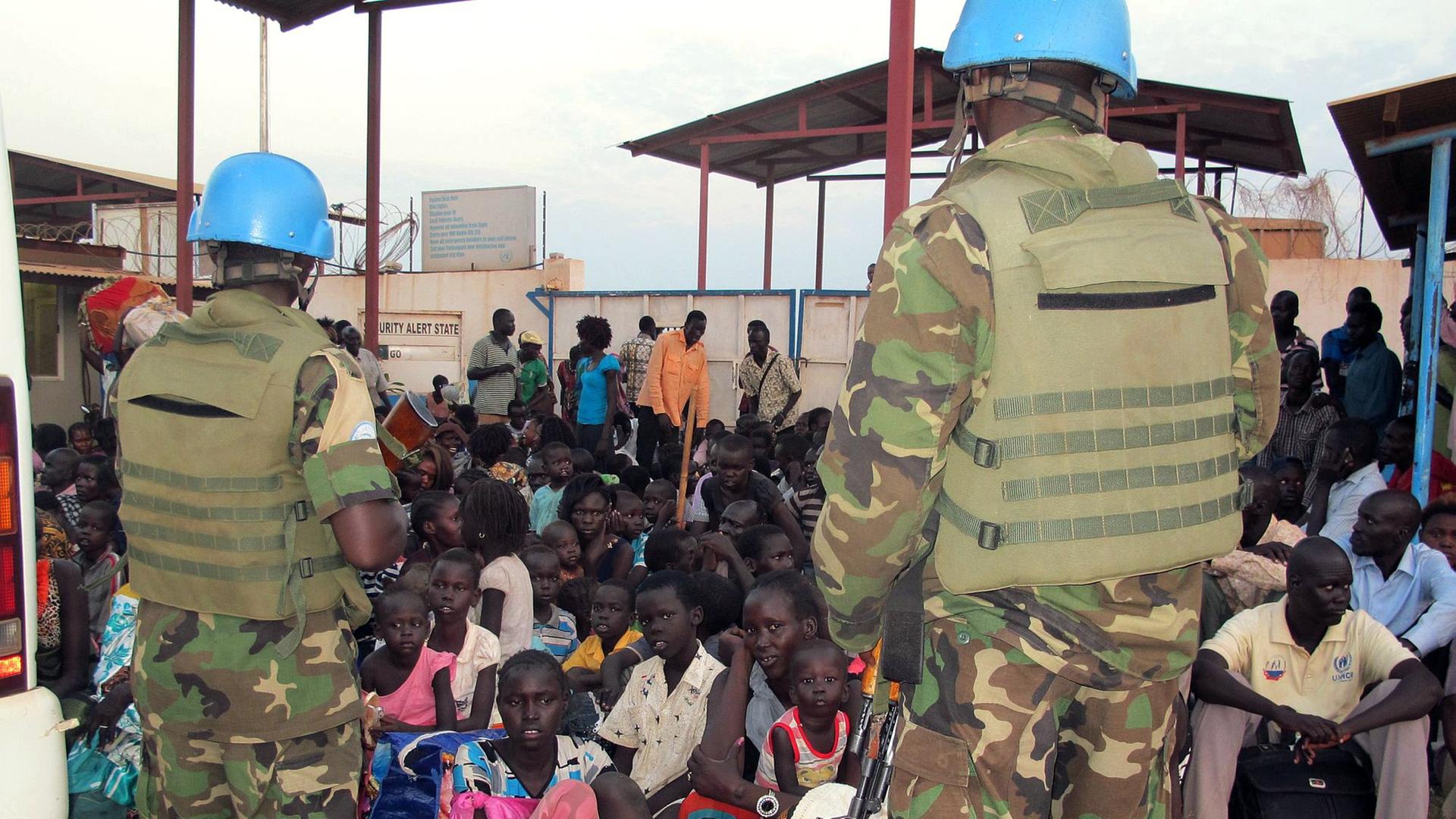 In einem UNO-Quartier in der südsudanesischen Hauptstadt Juba sitzen viele Flüchtlinge auf dem Boden, vor ihnen stehen zwei Blauhelmsoldaten der UNO.