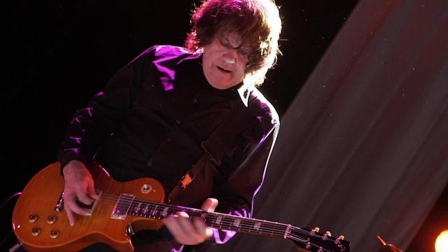 Ein Mann in dunkler Kleidung mit lockigen Haaren steht auf einer Bühne. Er hält eine Gitarre in der Hand.