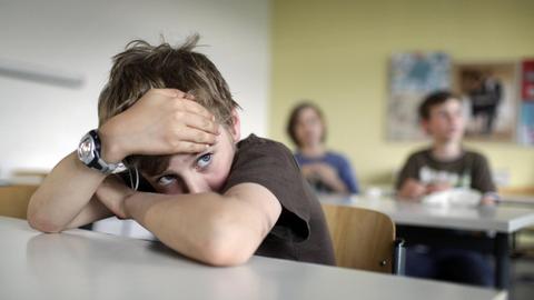 Ein Junge mit dunklem T-Shirt und zerzausten Haaren sitzt im Klassenzimmer und hat den Kopf auf die verschränkten Arme gestützt.