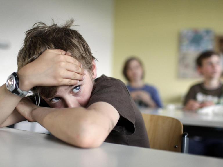  Ein Junge mit dunklem T-Shirt und zerzausten Haaren sitzt im Klassenzimmer und hat den Kopf auf die verschränkten Arme gestützt.