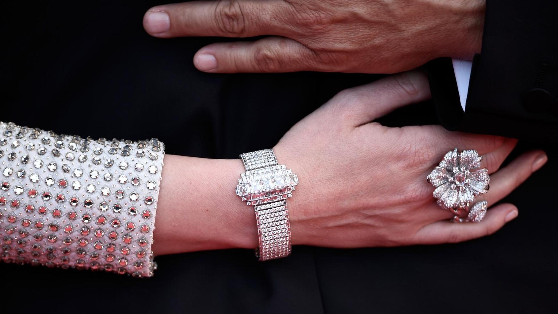 Eine Frauenhand mit grossem Diamantenring und-armband und silber glitzerndem Ärmel. Darüber eine Männerhand. Eine Frauenhand mit grossem Diamantenring und-armband und silber glitzerndem Ärmel. Darüber eine Männerhand.