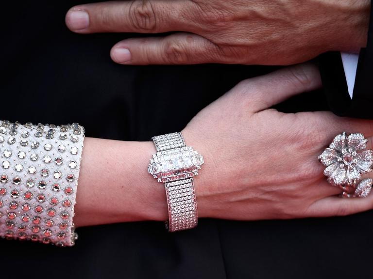 Eine Frauenhand mit grossem Diamantenring und-armband und silber glitzerndem Ärmel. Darüber eine Männerhand.