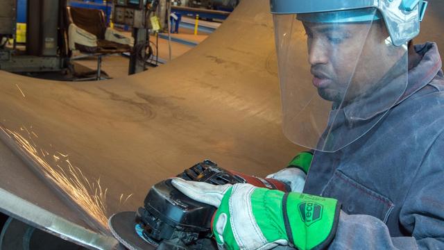 Der Arbeiter hat einen blauen Schutzhelm mit Sichtschutz auf und flext den Rand einer Metallröhre.