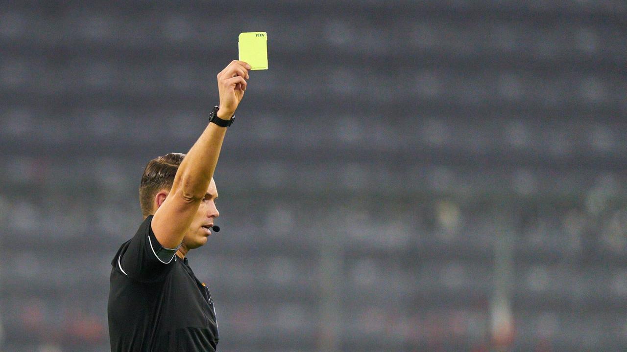 Ein schwarz gekleideter Fußballschiedsrichter zeigt mit erhobener rechter Hand die Gelbe Karte. Ein schwarz gekleideter Fußballschiedsrichter zeigt mit erhobener rechter Hand die Gelbe Karte.