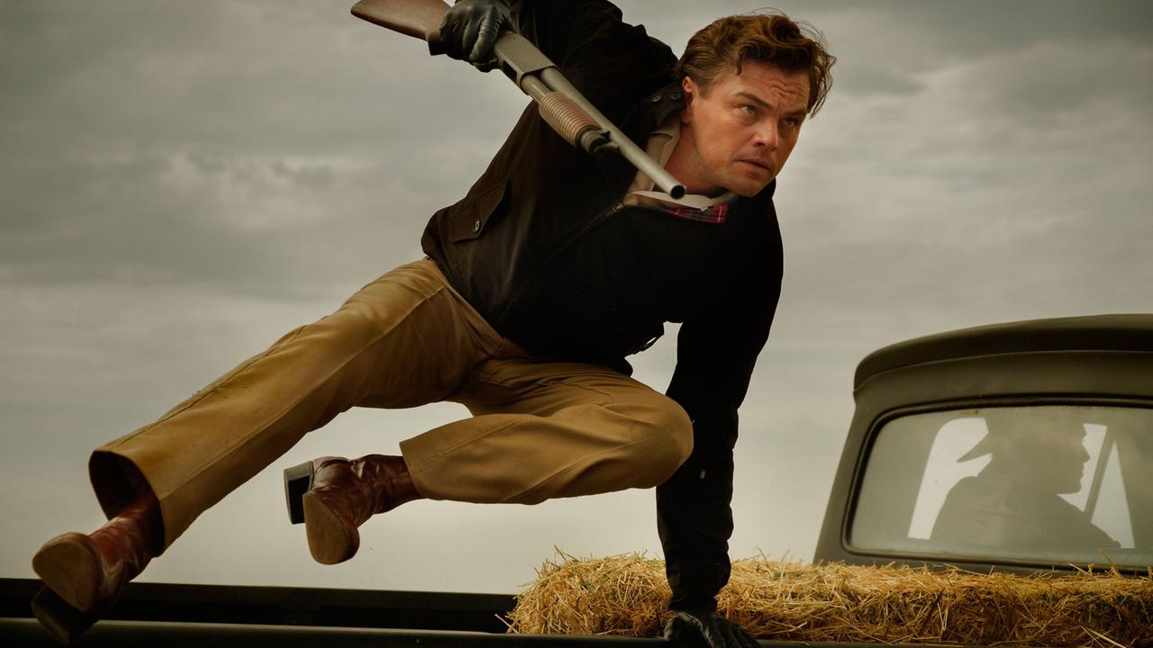 Das Bild zeigt eine Szene aus dem Tarantino-Film "Once upon a time in Hollywood". Der Schauspieler Leonardo DiCaprio springt darin mit einem Gewehr in der rechten Hand über die Ladefläche eines Pickups.