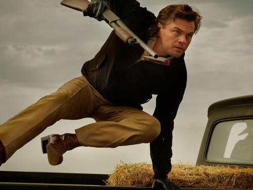 Das Bild zeigt den Schauspieler Leoonardo DiCaprio beim Sprung über ein Auto mit einem Gewehr in der Hand.