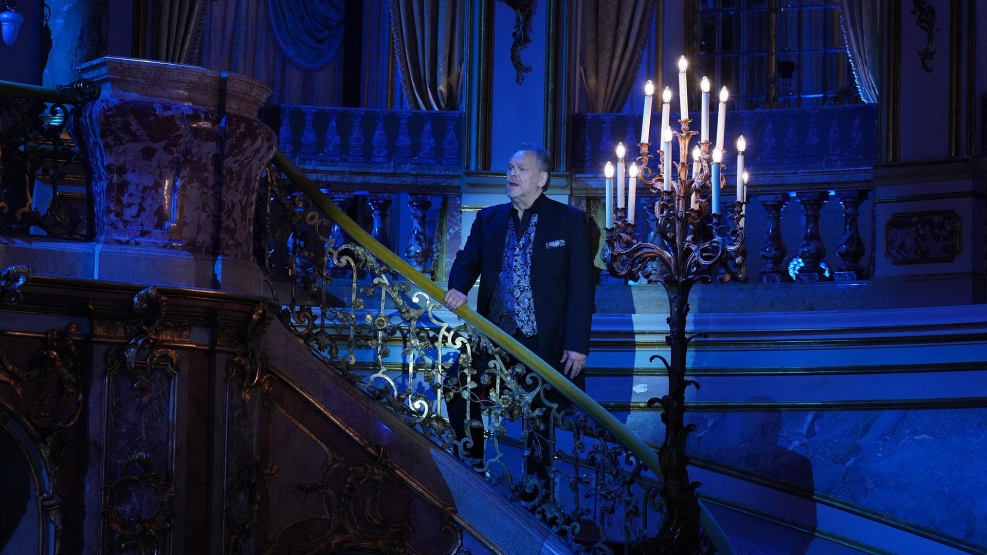 Ein Sänger steht auf einer barocken Treppe, direkt neben ihm ein Kronleuchter mit brennenden Kerzen. Alles ist in bläuliches Licht getaucht.