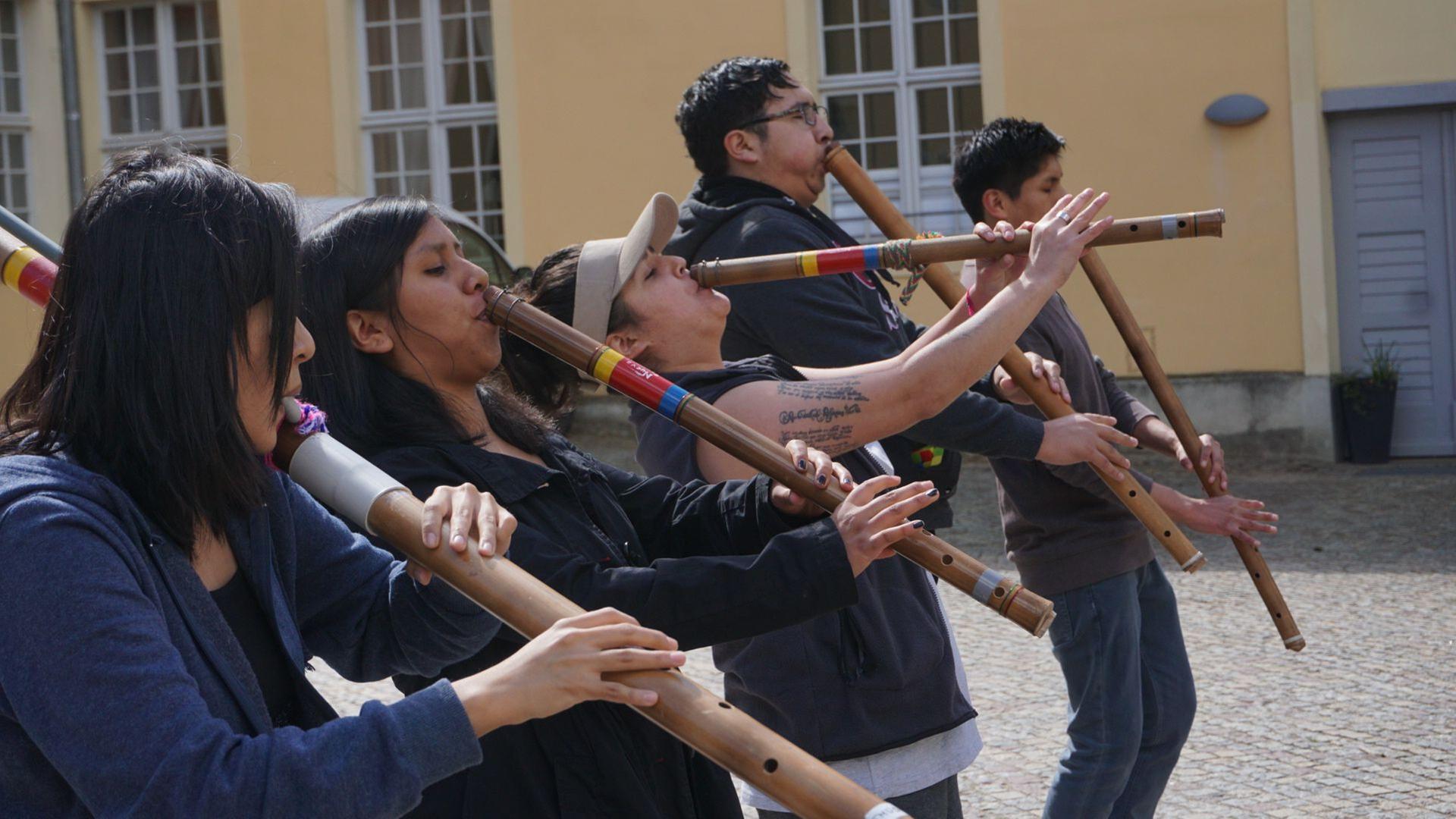 Drei Fraun und zwei Männer stehen nebeneinander und spielen auf indigenen Blas-Instrumenten. Drei Fraun und zwei Männer stehen nebeneinander und spielen auf indigenen Blas-Instrumenten.