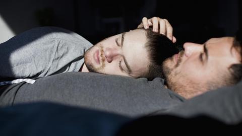 Ein schwules Paar liegt mit geschlossenen Augen im Bett und die Sonne scheint ihnen ins Gesicht.