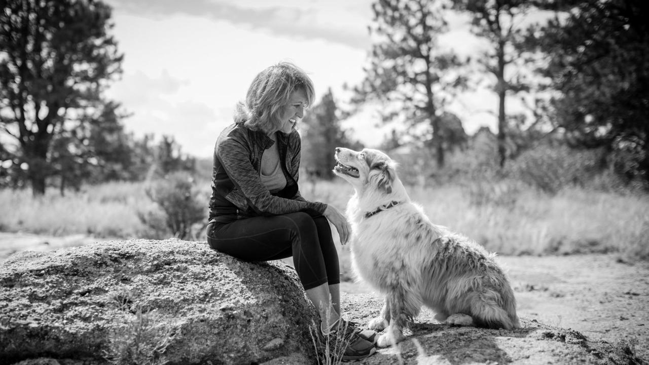 Eine Frau sitzt auf einem Stein und lächelt ihren Hund an, der zu ihr aufschaut. Eine Frau sitzt auf einem Stein und lächelt ihren Hund an, der zu ihr aufschaut.