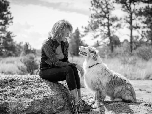 Eine Frau sitzt auf einem Stein und lächelt ihren Hund an, der zu ihr aufschaut.