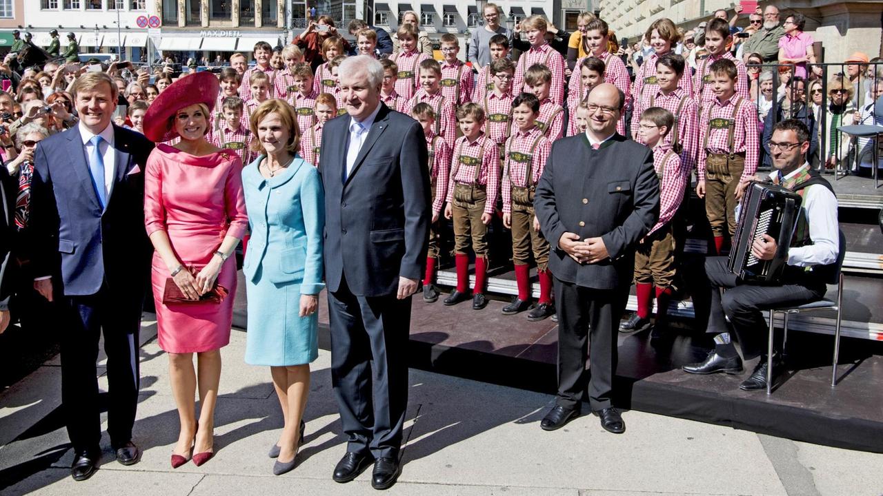 Horst Seehofer steht neben Königin Maxima und Willem-Alexander, hinter ihnen der Tölzer Knabenchor auf einer Bühne in rot-weiß karierten Hemden und Lederhosen.
