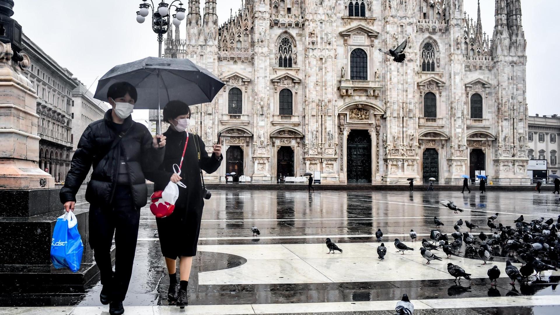 Touristen gehen mit Mundschutz bekleidet und einem Regenschirm über die Piazza del Duomo vor dem Mailänder Dom. Touristen gehen mit Mundschutz bekleidet und einem Regenschirm über die Piazza del Duomo vor dem Mailänder Dom.
