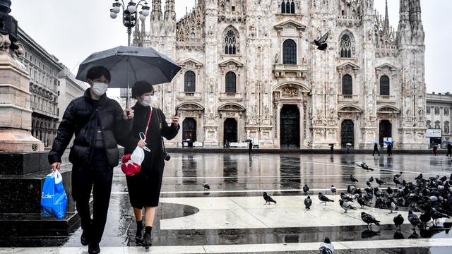 Touristen gehen mit Mundschutz bekleidet und einem Regenschirm über die Piazza del Duomo vor dem Mailänder Dom.