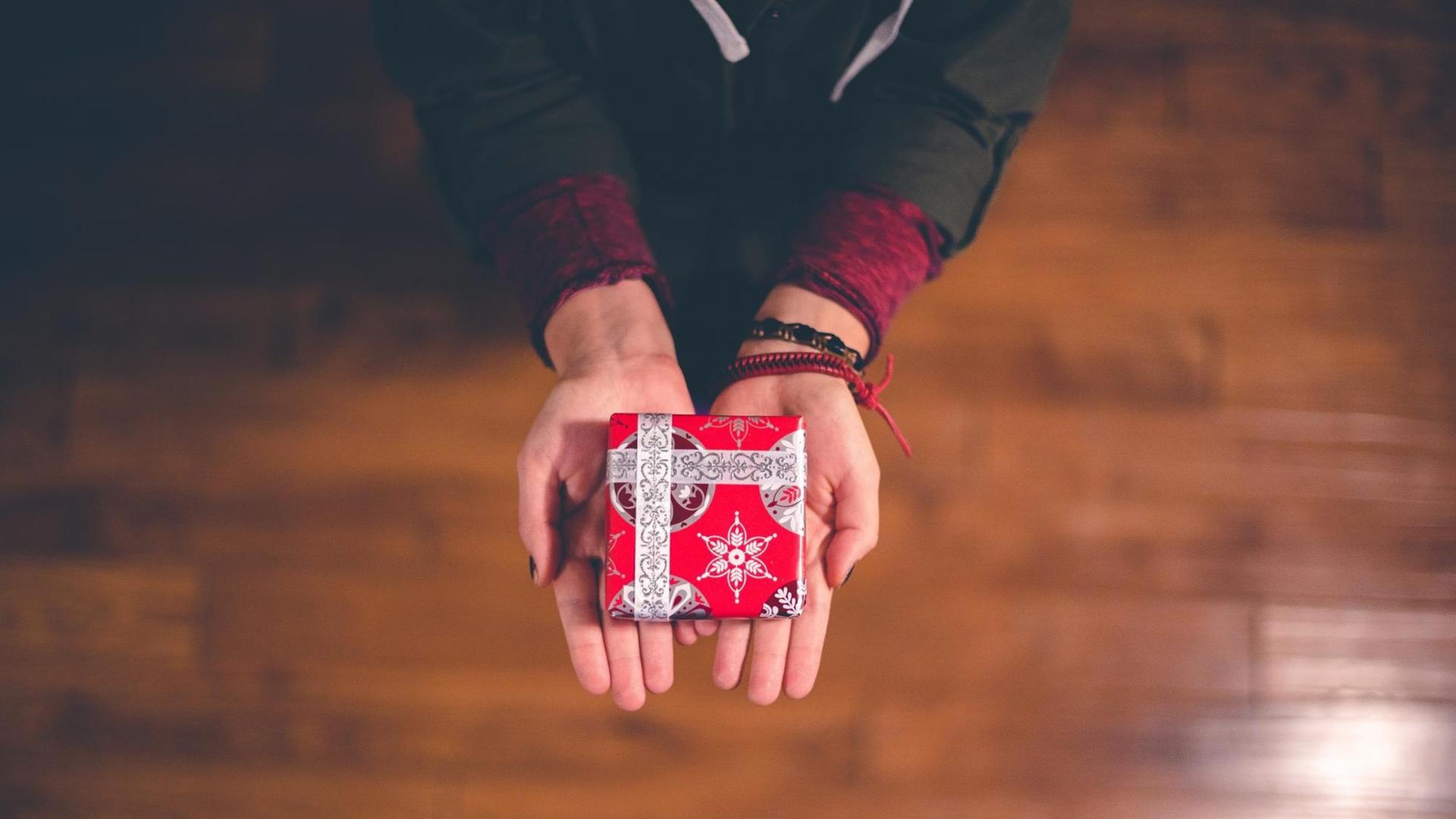 Ein kleines Geschenk in weihnachtlicher Verpackung wird auf zwei Händen überreicht.