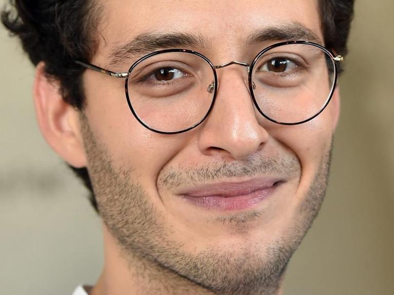 Ein junger Mann mit kurzen dunklen Haare und Brille lächelt in die Kamera. 