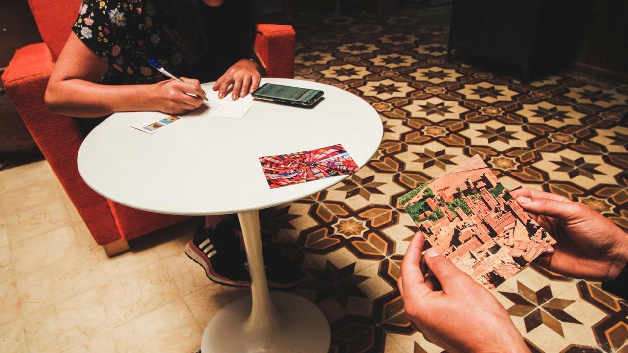 Eine Frau sitzt an einem Tisch und schreibt eine Postkarte. Eine Person daneben schaut sich eine Postkarte an. Eine Frau sitzt an einem Tisch und schreibt eine Postkarte. Eine Person daneben schaut sich eine Postkarte an.