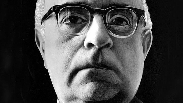 Ein Nahaufnahme von Theodor W. Adorno vor schwarzem Hintergrund. Er trägt eine Hornbrille und schaut aufmerksam dem Betrachter entgegen. Ein Nahaufnahme von Theodor W. Adorno vor schwarzem Hintergrund. Er trägt eine Hornbrille und schaut aufmerksam dem Betrachter entgegen.