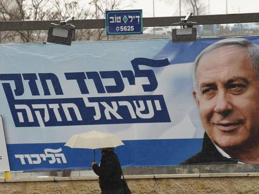 Eine Frau mit Regenschirm läuf an einem großen, blauweißen Wahlplakat vorbei, das Benjamin Netanjahu in selbstbewusster Pose zeigt.