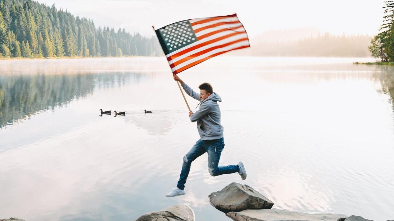 Ein junger Mann springt locker über Steine, die aus einem Gewässer ragen, während er stolz die Flagge der Vereinigten Staaten von Amerika wedelt. Im Hintergrund sieht man im Nebel versunkenes Waldgebirge. Ein junger Mann springt locker über Steine, die aus einem Gewässer ragen, während er stolz die Flagge der Vereinigten Staaten von Amerika wedelt. Im Hintergrund sieht man im Nebel versunkenes Waldgebirge.