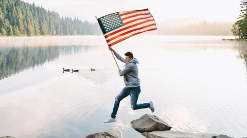 Ein junger Mann springt locker über Steine, die aus einem Gewässer ragen, während er stolz die Flagge der Vereinigten Staaten von Amerika wedelt. Im Hintergrund sieht man im Nebel versunkenes Waldgebirge.