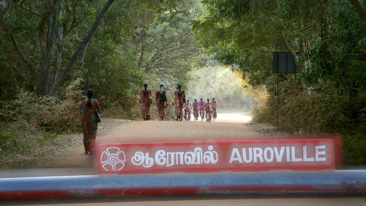 Inderinnen mit Saris auf Fahrrädern laufen einen Weg entlang, im Vordergrund der Schriftzug "Auroville"