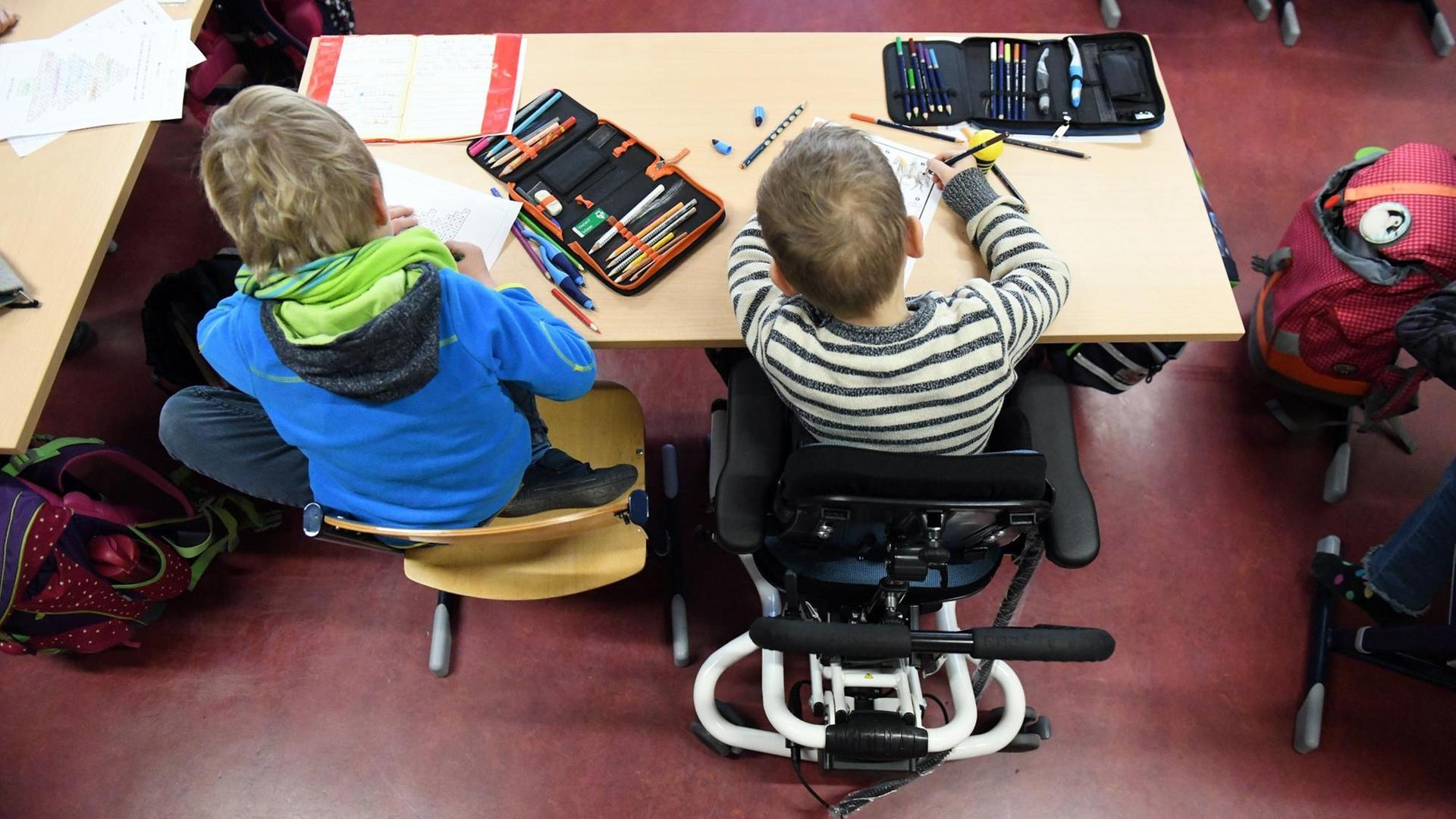 Ein Kind im Rollstuhl sitzt mit einem zweiten Kind an einem Schultisch. Ein Kind im Rollstuhl sitzt mit einem zweiten Kind an einem Schultisch.