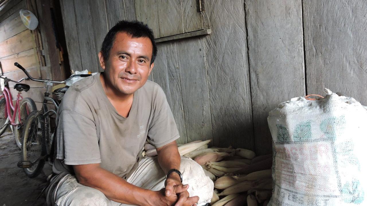 Ein Mann sitzt in beigem T-Shirt und mit gefalteten Händen vor einem Sack mit Kakaopflanzen.