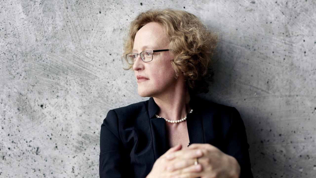 Eine Frau mit schulterlangem, blonden, lockigem Haar sitzt vor einer grauen Betonwand und schaut konzentriert nach links. Eine Frau mit schulterlangem, blonden, lockigem Haar sitzt vor einer grauen Betonwand und schaut konzentriert nach links.