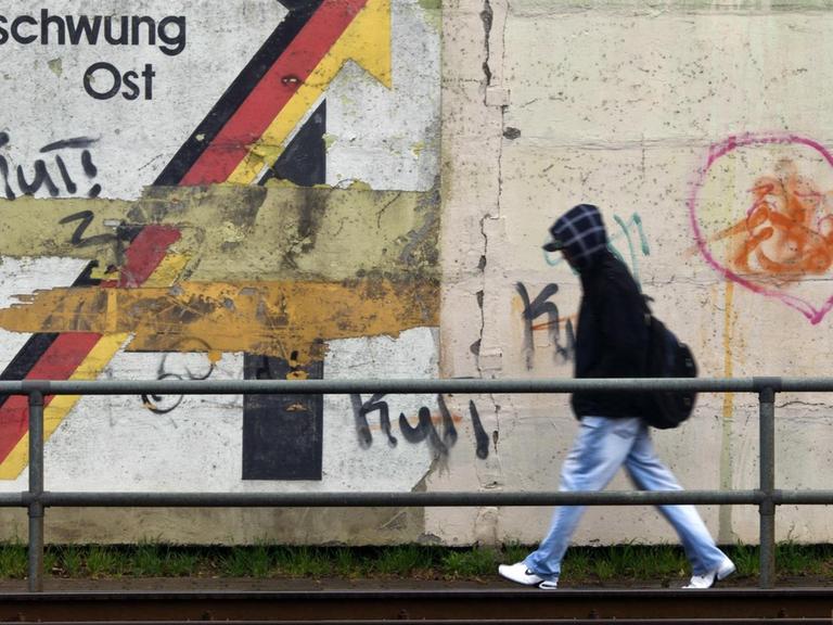 Ein Passant geht am 30.03.2012 an einer Schallschutzmauer in Magdeburg an einem aus der Wendezeit stammenden verwitterten Wandbild mit dem Schriftzug «Gemeinschaftswerk Aufschwung Ost» vorbei.