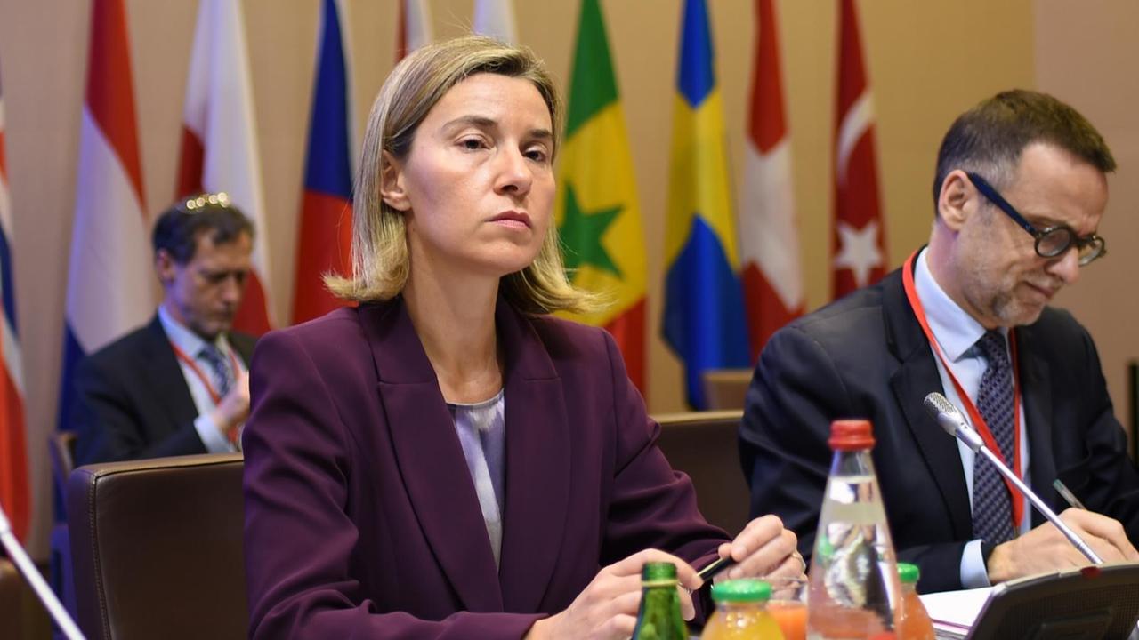 Mogherini sitzt im lila Sakko mit ernstem Blick an einem Tisch; neben ihr schreibt ein weiterer Teilnehmer. Im Hintergrund stehen Fahnen der beteiligten Länder.
