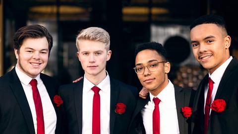 Vier junge Männer in schwarzen Anzügen, roten Schlips und jeweils einer roten Rose am Revers lächeln eng beieinander stehend in die Kamera.