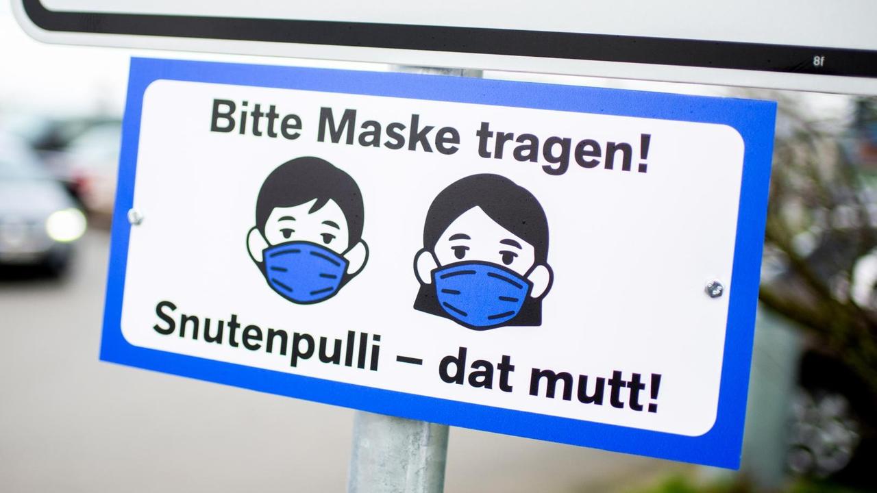 Ein Schild mit der Aufschrift „Bitte Maske tragen!“ und dem plattdeutschen Zusatz „Snutenpulli - dat mutt!“ hängt an der Zufahrt zu einem Parkplatz.