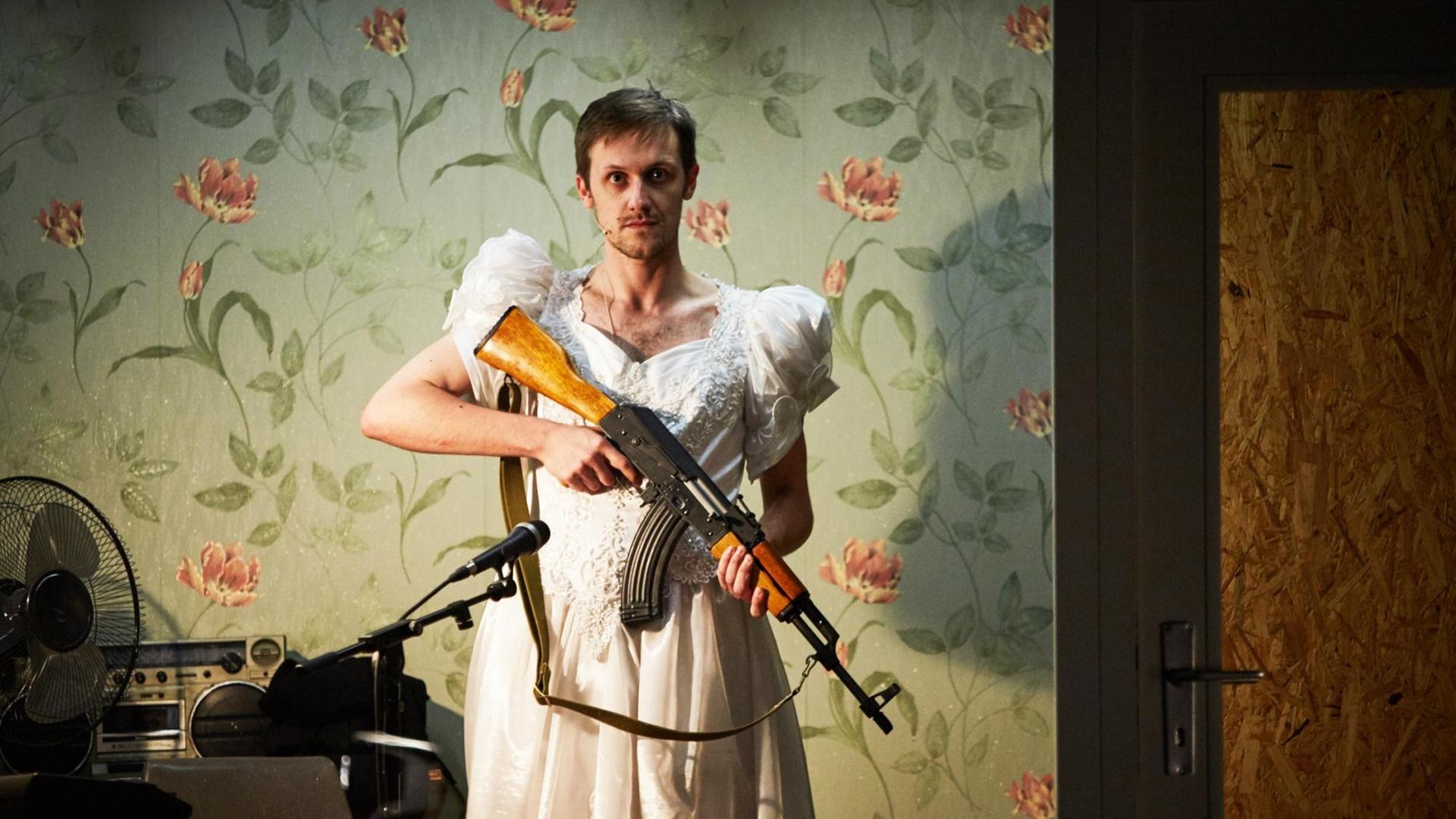 Ein Schauspieler im Brautkleid und mit einer Kalaschnikov in im Anschlag. Im Hintergrund grüne Blümchentapete. Ein Schauspieler im Brautkleid und mit einer Kalaschnikov in im Anschlag. Im Hintergrund grüne Blümchentapete.