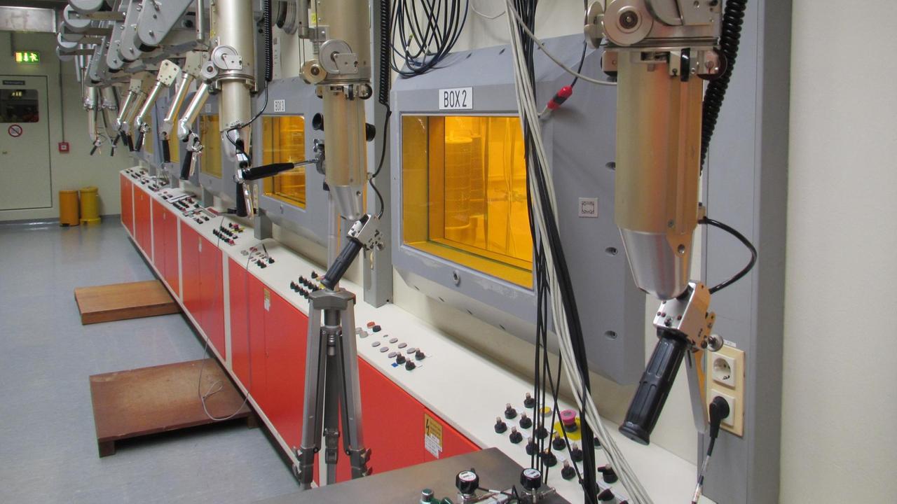 Blick auf die Reihe der "heißen" Versuchsboxen im Strahlenlabor des KIT: Vor jeder der gelb scheinenden Boxen hängen zwei Griffarme aus Metall, mit denen die Arm- und Handbewegungen des Operators auf die Griffarme im Inneren der Box übertragen werden Blick auf die Reihe der "heißen" Versuchsboxen im Strahlenlabor des KIT: Vor jeder der gelb scheinenden Boxen hängen zwei Griffarme aus Metall, mit denen die Arm- und Handbewegungen des Operators auf die Griffarme im Inneren der Box übertragen werden