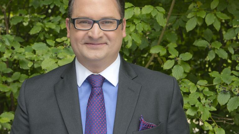 Ein Mann in Geschäftskleidung und Brille steht vor einer grünen Hecke und lächelt. Ein Mann in Geschäftskleidung und Brille steht vor einer grünen Hecke und lächelt.