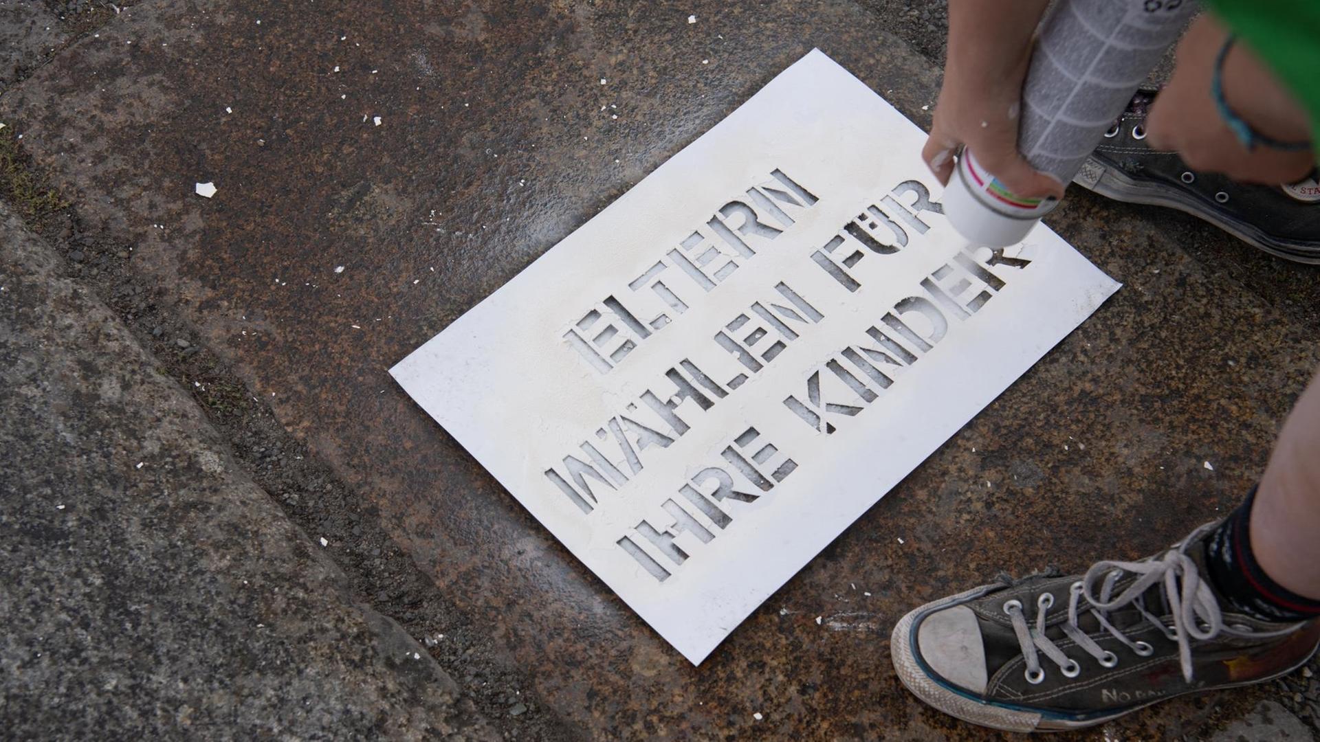 Jemand schreibt mit weißer Farbe und einer Schablone die Worte "Eltern wählen für ihre Kinder" auf den Boden. Jemand schreibt mit weißer Farbe und einer Schablone die Worte "Eltern wählen für ihre Kinder" auf den Boden.