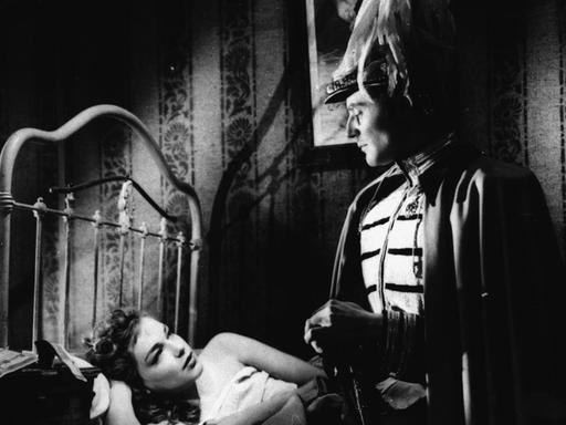 Ein schwarzweißes Filmstill aus Max Ophuels' Film "Der Reigen" (La Ronde) von 1950 zeigt eine Szene mit Gerard Philippe und Simone Signoret, ihn in Uniform stehend dieauf dem Bett liegende schmachtend anblickend