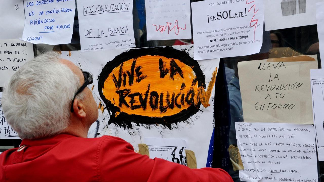 Ein älterer Mann steht vor einer Plakatwand, auf der die Aufschrift "Vive La Revolucia" zu lesen ist.