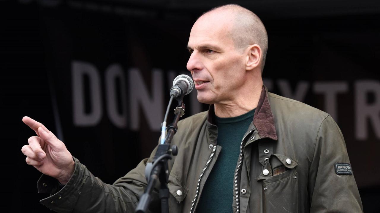 Janis Varoufakis, mit kahlgeschorenem Kopf und brauner Jacke spricht in ein Mikrofon und gestikuliert dabei mit der rechten Hand. Janis Varoufakis, mit kahlgeschorenem Kopf und brauner Jacke spricht in ein Mikrofon und gestikuliert dabei mit der rechten Hand.