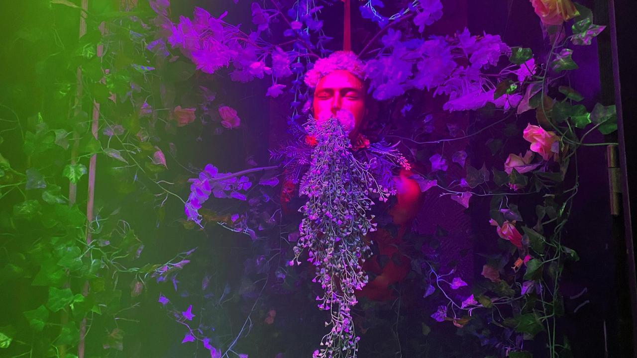 Ausstellungsobjekt einer Figur mit Maske, umringt von künstlichen Pflanzen, im farbigen Licht.