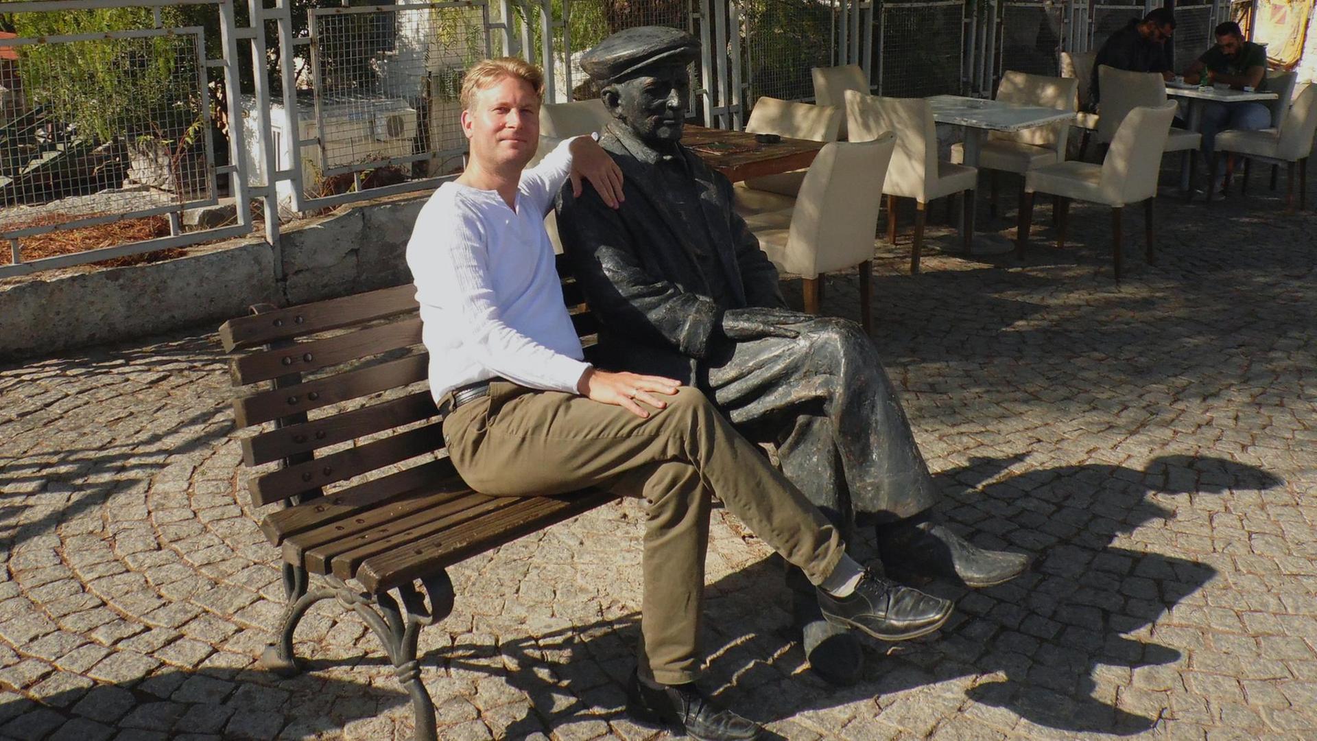 Ein blonder Mann sitzt neben einer männlichen Statue auf einer Parkbank. Beide haben die Beine überschlagen.