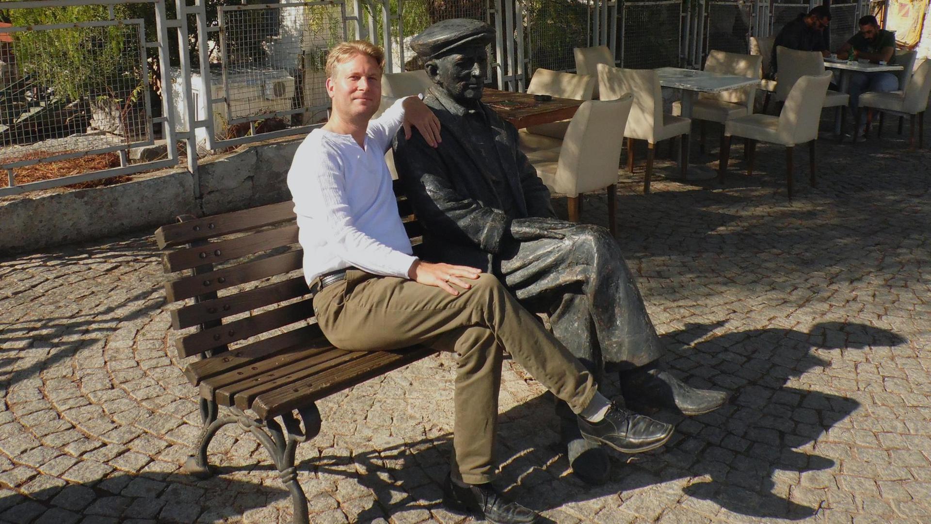 Ein blonder Mann sitzt neben einer männlichen Statue auf einer Parkbank. Beide haben die Beine überschlagen. Ein blonder Mann sitzt neben einer männlichen Statue auf einer Parkbank. Beide haben die Beine überschlagen.
