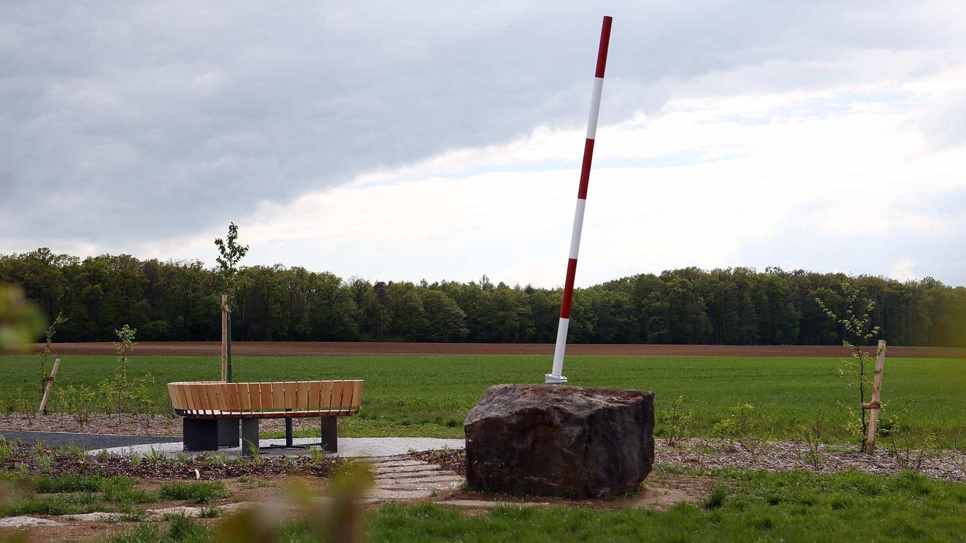 Auf einem Acker steht ein Denkmal mit einem großen Stein, von dem ein rot-weißer Stab schräg aufragt, daneben eine halbrunde Sitzbank und frisch gepflanzte Bäume. Auf einem Acker steht ein Denkmal mit einem großen Stein, von dem ein rot-weißer Stab schräg aufragt, daneben eine halbrunde Sitzbank und frisch gepflanzte Bäume.