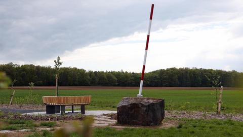 Auf einem Acker steht ein Denkmal mit einem großen Stein, von dem ein rot-weißer Stab schräg aufragt, daneben eine halbrunde Sitzbank und frisch gepflanzte Bäume.
