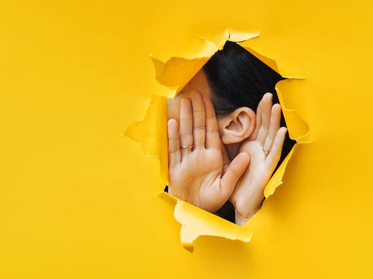 In eine gelbe Pappe ist ein Loch gerissen, durch das man ein Ohr und zwei Hände, die zum Zuhören aufgestellt sind, erkennt.